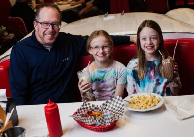 A father and his two young daughters smiling at a diner booth, with milkshakes and a basket of food on the table