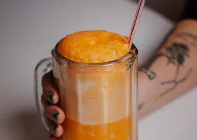 A hand holding a mug filled with an orange and white frothy beverage (likely an ice cream float or creamsicle shake) with a straw