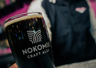 A man holding a glass of dark Nokomis Craft Ales beer with a blurred pink background