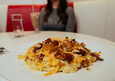 A plate of macaroni and cheese topped with bacon, shredded cheese, with a woman and a milkshake blurred in the background