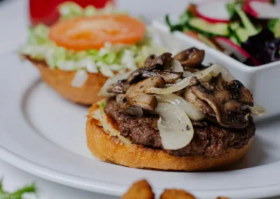 A plate with an open-faced hamburger topped with sauteed mushrooms and onions, a side salad, and fried appetizers in the foreground