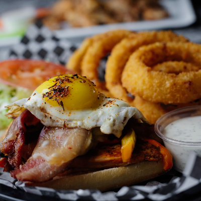 A savory dish featuring a toasted bun topped with bacon, cheese, a fried egg, and fresh veggies, accompanied by crispy onion rings and ranch dip