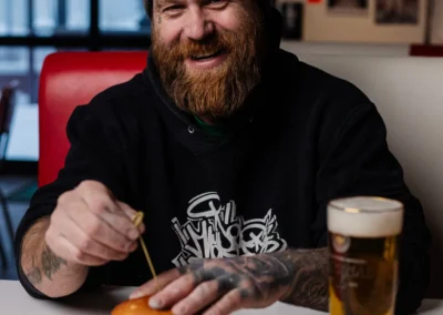 A smiling man with a beard and tattoos wearing a beanie, sitting in a diner booth with a burger and a glass of beer