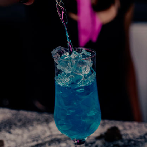 A vibrant blue cocktail with ice is being poured, set against a blurred background featuring a person in a black shirt