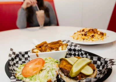 An open-face cheeseburger topped with pickles on a bun next to a bowl of poutine, with a woman drinking a milkshake