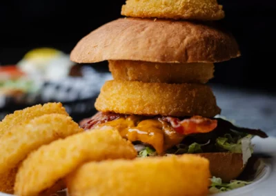 Tall cheeseburger stacked with bacon, melted cheese and a small fried round on a skewer, served with a side of onion rings