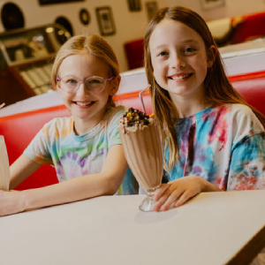 Two smiling young girls in tie-dye shirts sitting in a diner booth with a large chocolate milkshake topped with sprinkles between them
