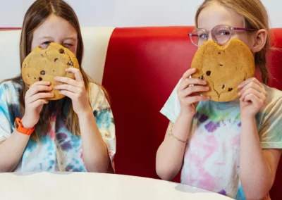 Two young girls in tie-dye shirts sitting in a diner booth, each covering their mouth with a large chocolate chip cookie